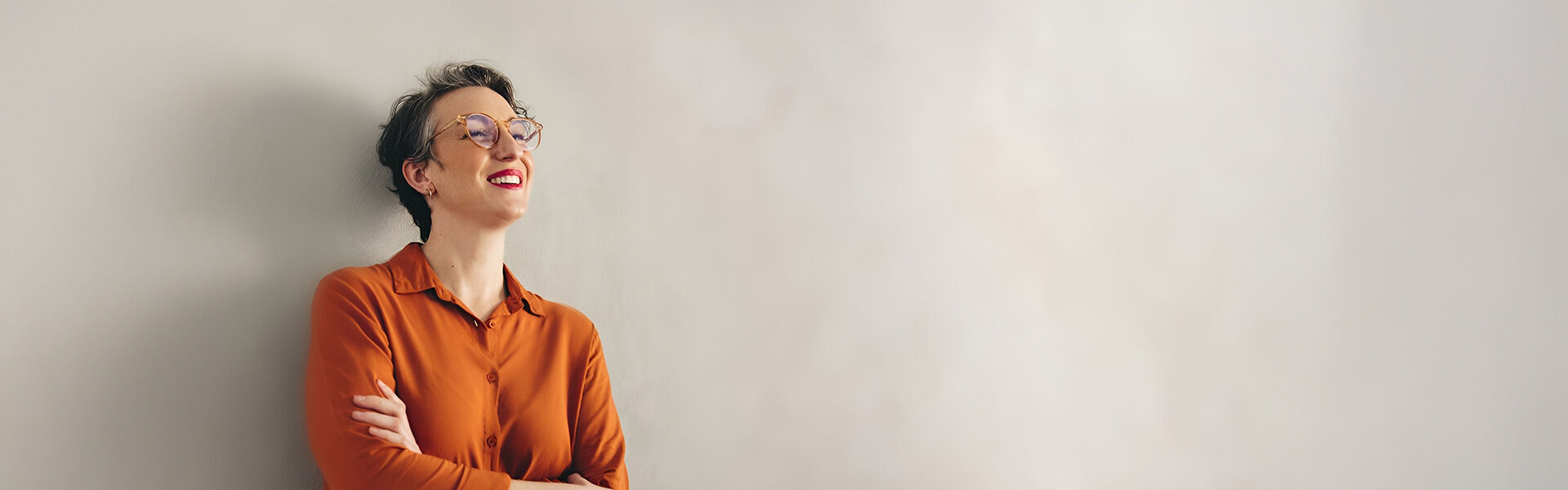 eine Frau in einem orangefarbenen Hemd und einer Brille, die an eine Wand gelehnt ist