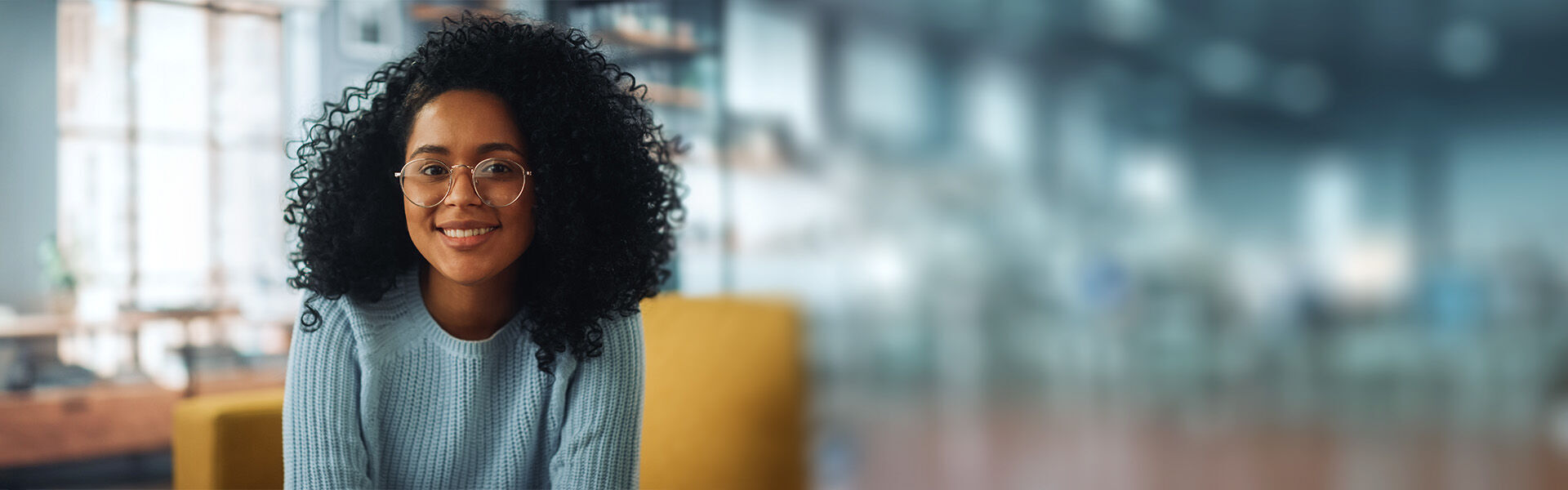 Junge Berufsanfängerin sitzt in einem undeutlich zu erkennenden Raum und schaut fröhlich und optimistisch nach vorne.